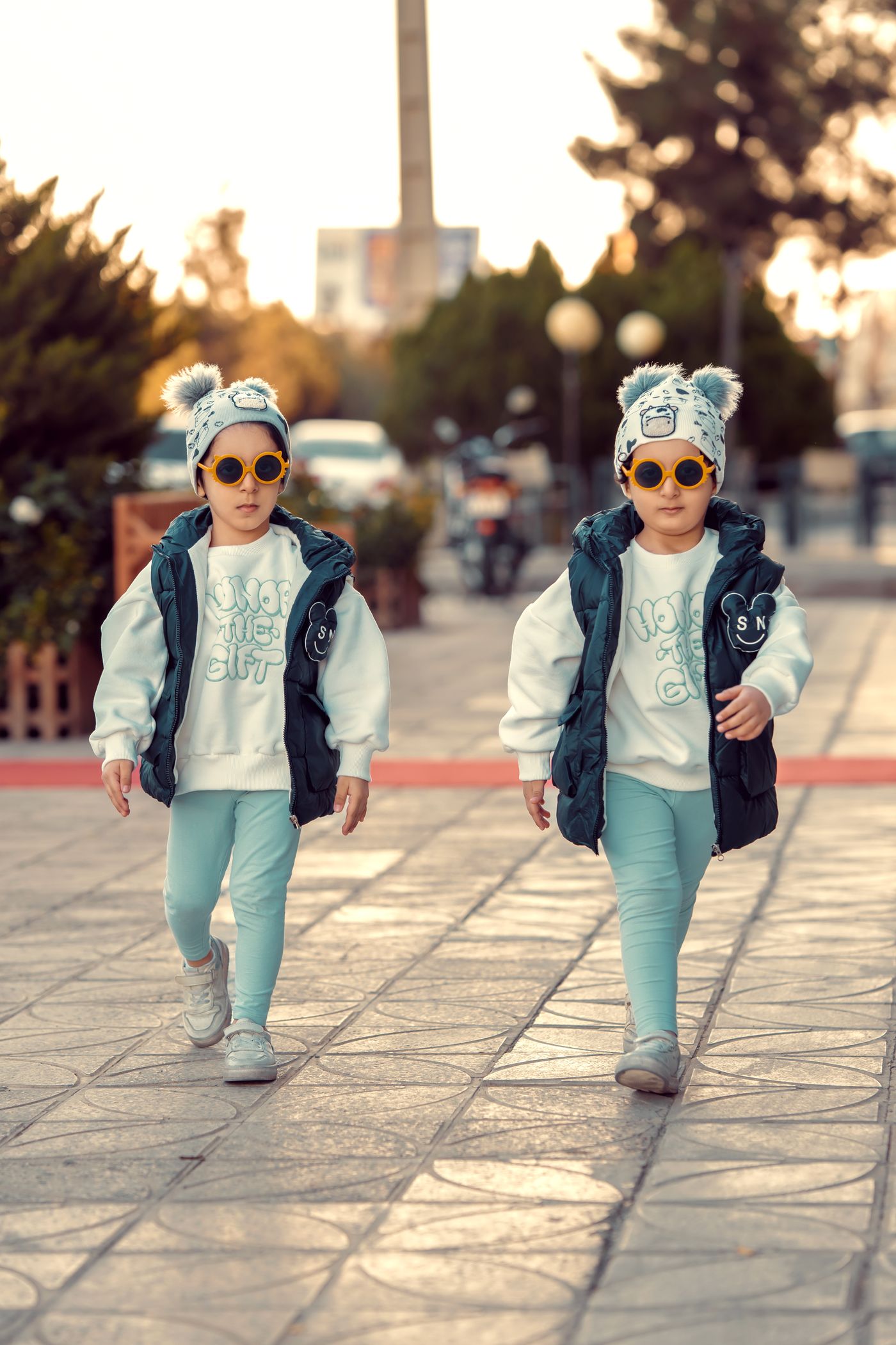 Two children wearing pale blue outfits, dark vests and knit hats.