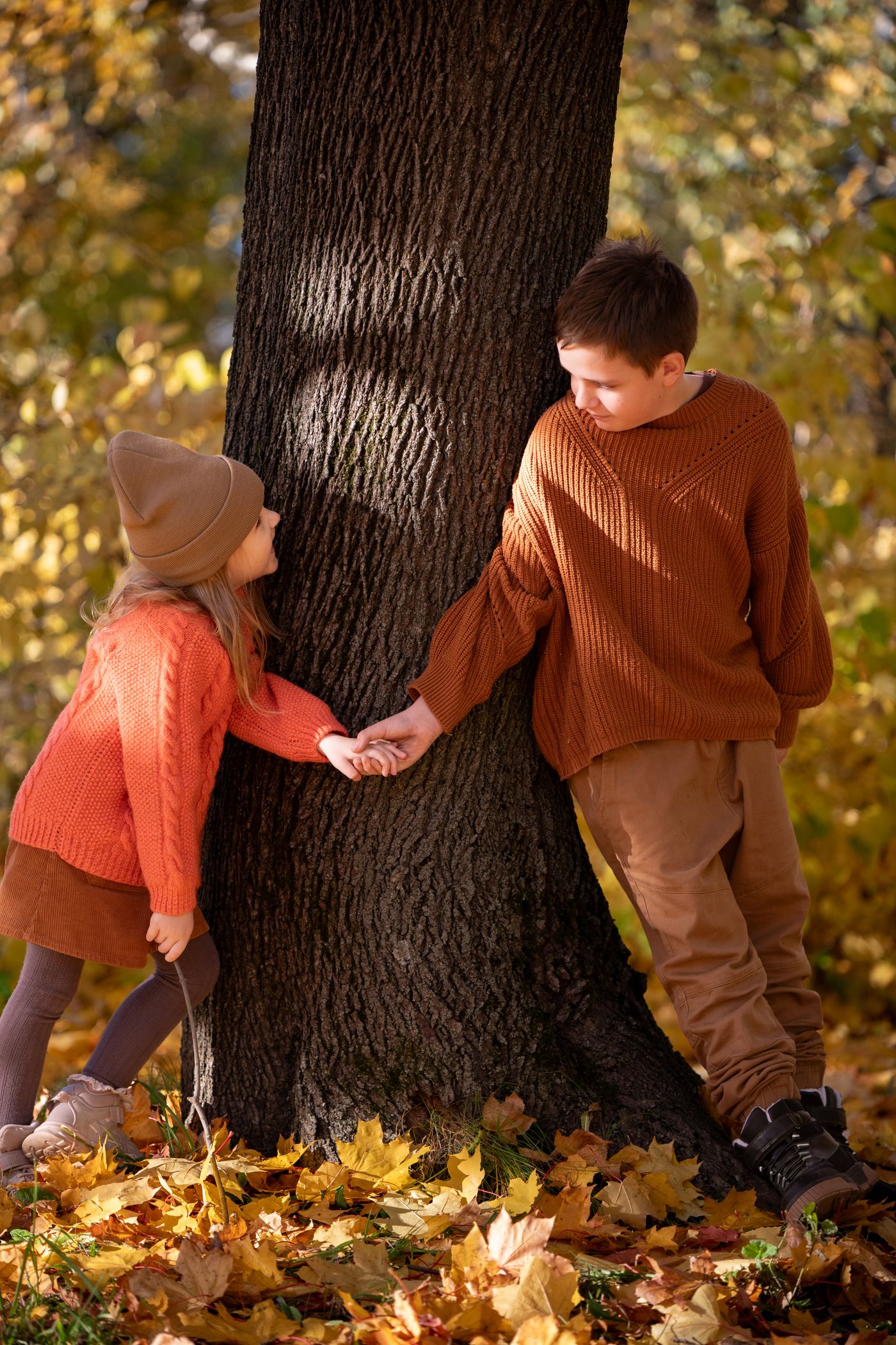 Two children wearing orange and brown sweaters beside a tree.