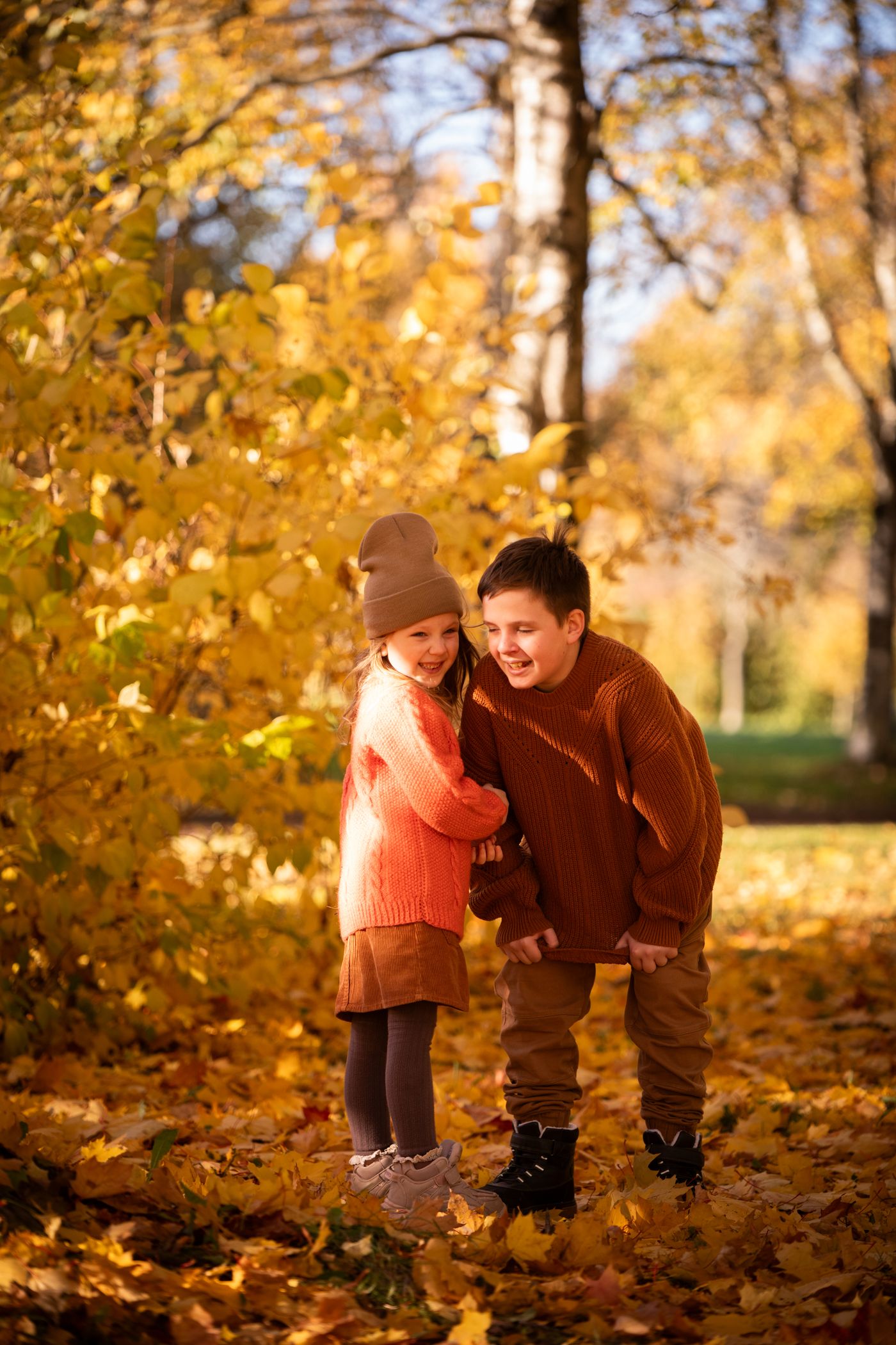 Two children in warm-colored sweaters standing among autumn leaves.